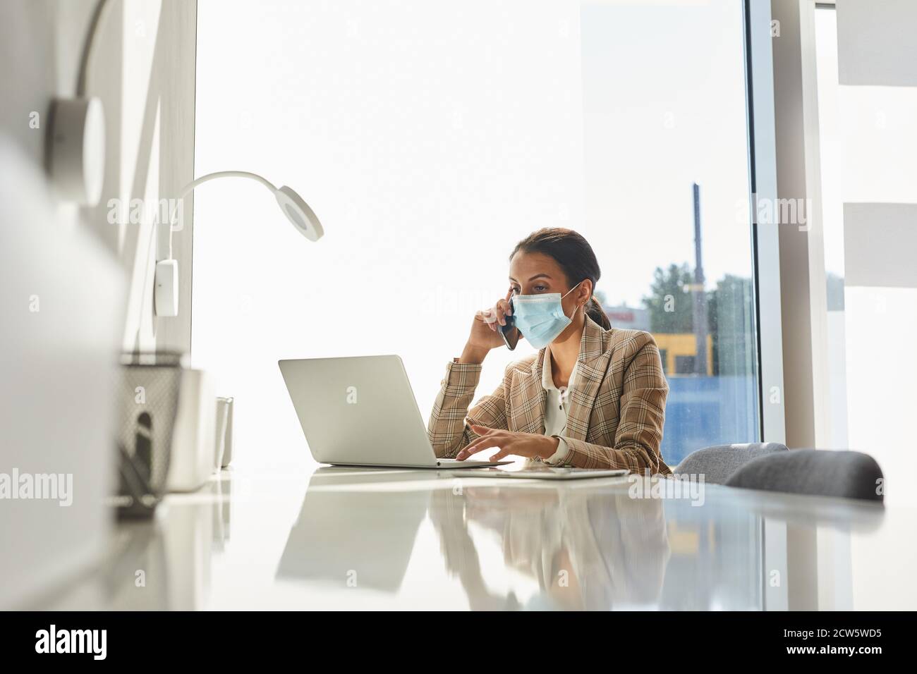 Portrait d'une femme d'affaires de race mixte portant un masque et parlant au smartphone tout en travaillant dans un bureau blanc, espace de copie Banque D'Images