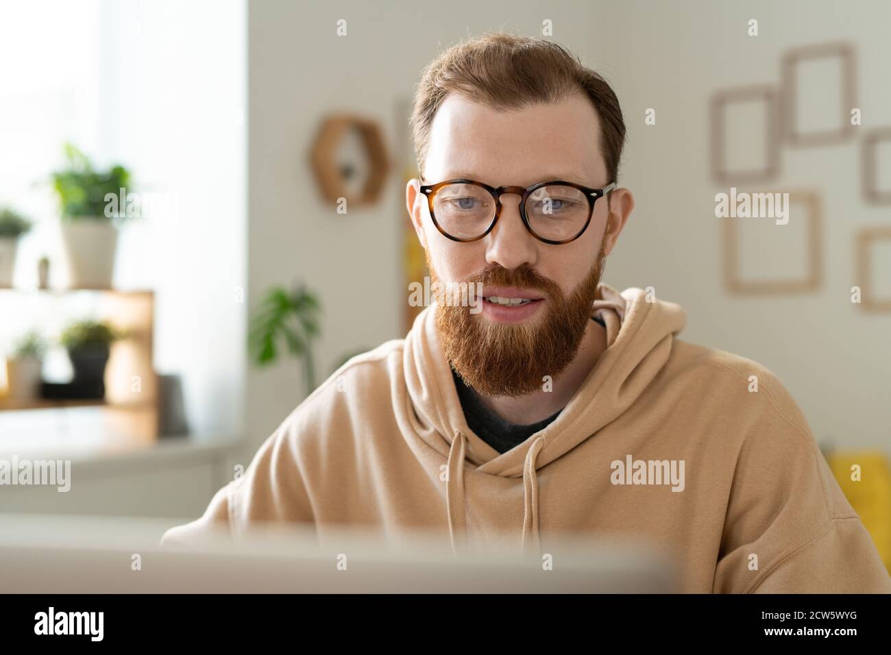 Jeune designer freelance barbu en lunettes regardant l'écran d'ordinateur portable Banque D'Images