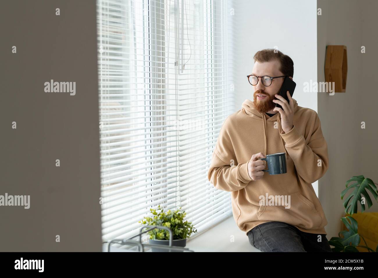 Jeune homme d'affaires à barbe en vêtements décontractés et lunettes de vue parlant sur smartphone Banque D'Images