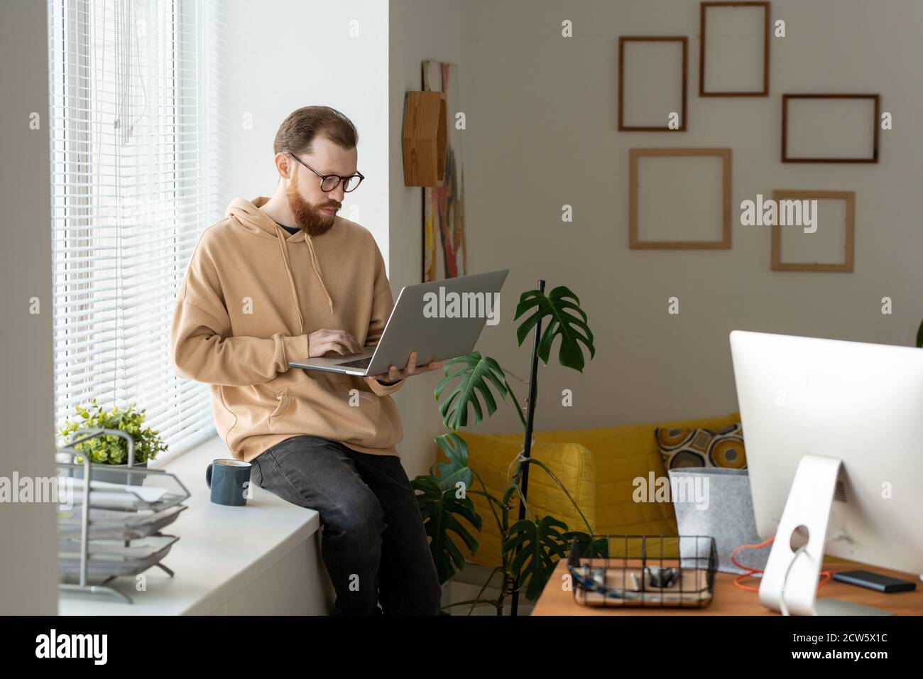 Jeune freelance barbu utilisant un ordinateur portable tout en réseau sur windowsill Banque D'Images