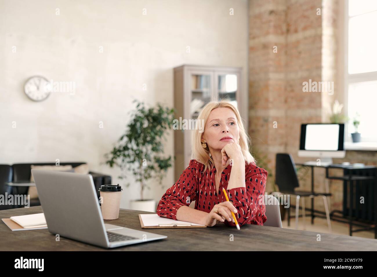 Femme d'affaires blonde et mûre pensive assise devant un ordinateur portable et contemplation Banque D'Images