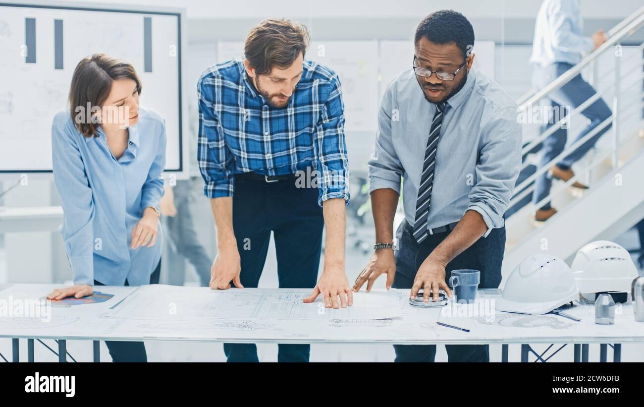 Dans le centre d'ingénierie industrielle : divers groupes d'ingénieurs et de techniciens réunis autour de la table démêle les feuilles de conception de moteur Banque D'Images