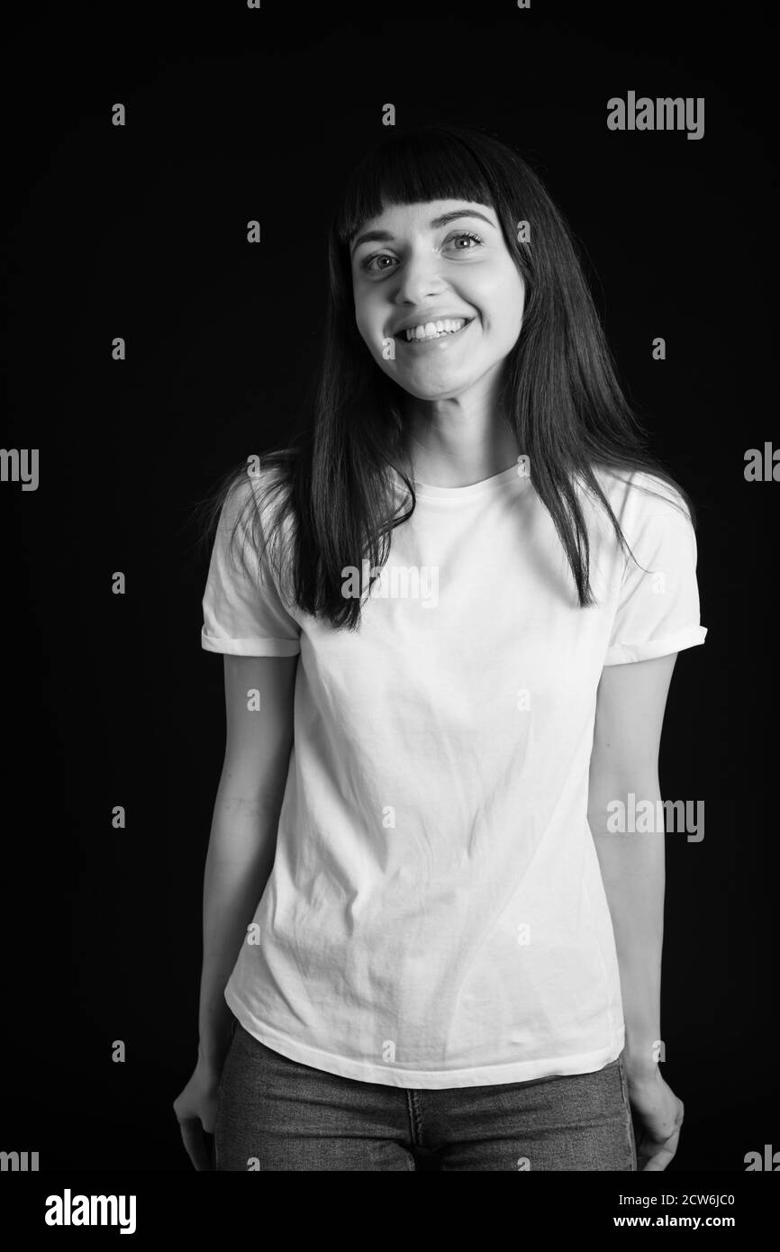 Portrait studio d'une jolie femme brune dans un t-shirt blanc, debout, souriant sur un fond noir Uni, regardant l'appareil photo Banque D'Images