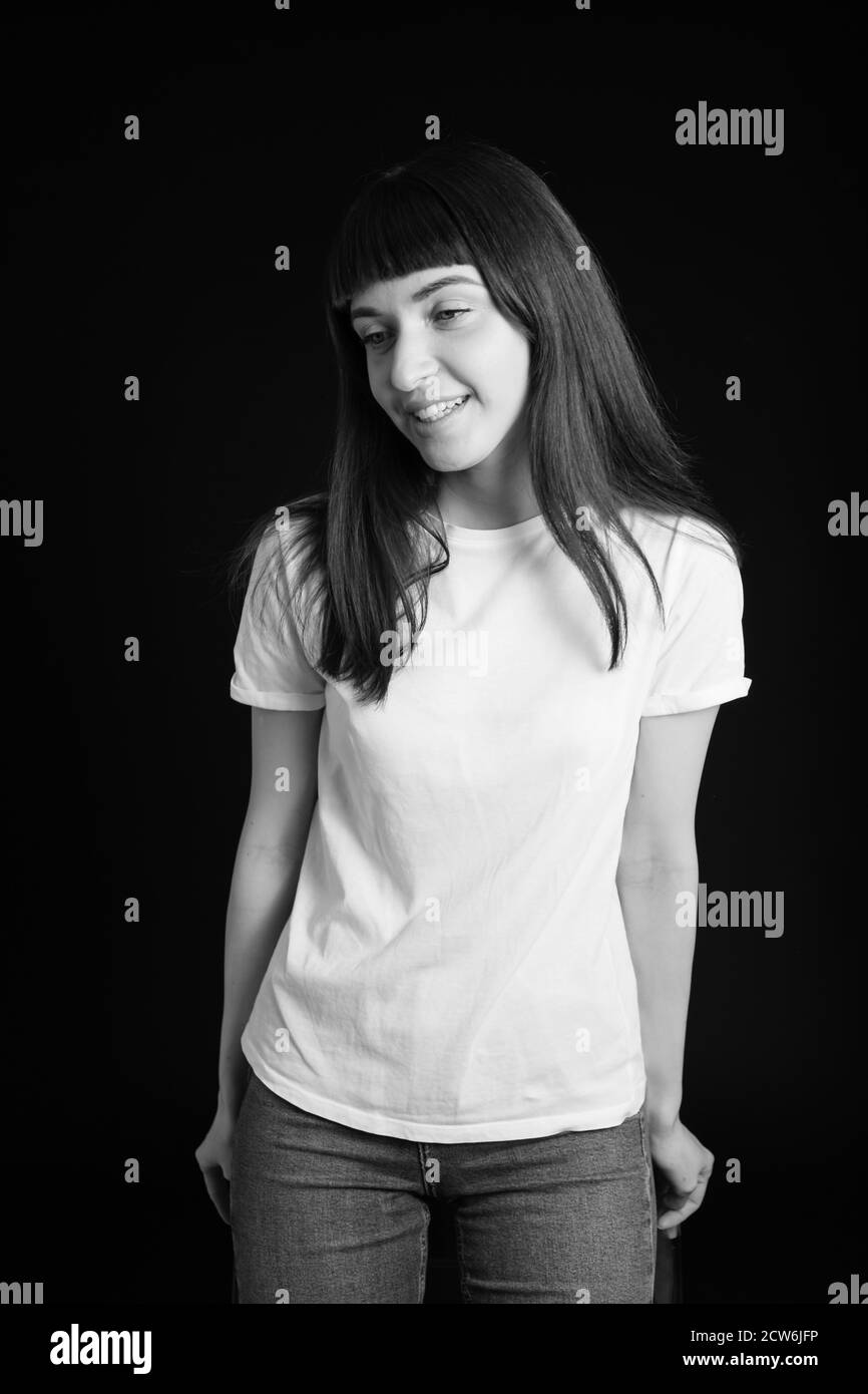 Portrait studio d'une jolie femme brune dans un t-shirt blanc, debout, souriant sur un fond noir Uni, regardant l'appareil photo Banque D'Images