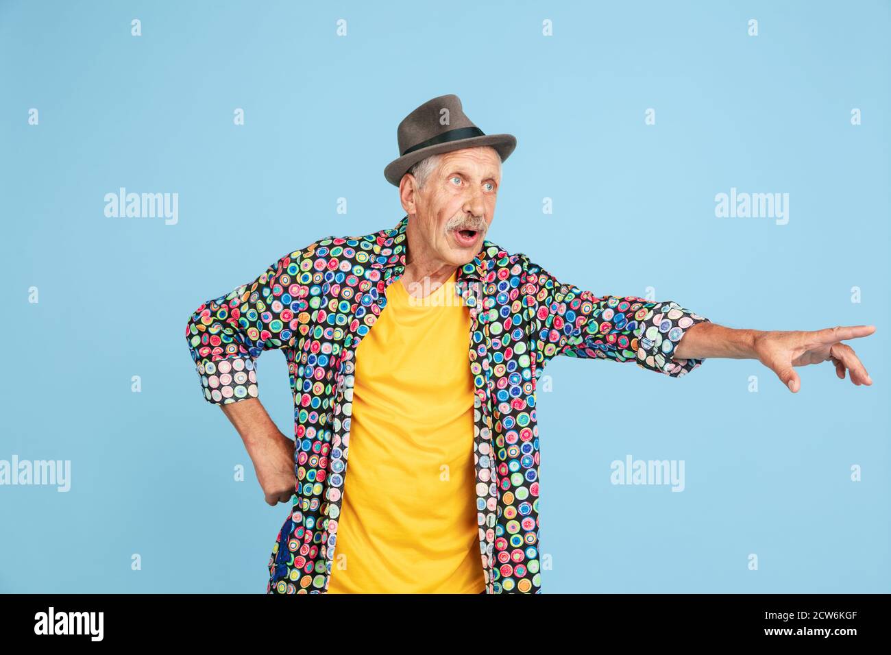 Choqué de pointer, de choisir. Portrait de l'homme hipster senior dans un chapeau élégant isolé sur fond bleu studio. Concept de mode de vie technologique et joyeux pour les personnes âgées. Couleurs tendance, toujours jeune. CopySpace pour publicité. Banque D'Images