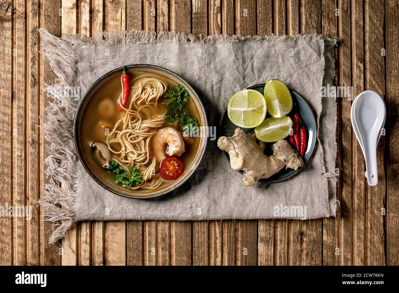 Soupe thaïlandaise épicée traditionnelle tom yum kung avec champignons shiitake et crevettes, ingrédients en plaque, cuillère sur tissu de lin sur fond de planche en bois. Banque D'Images