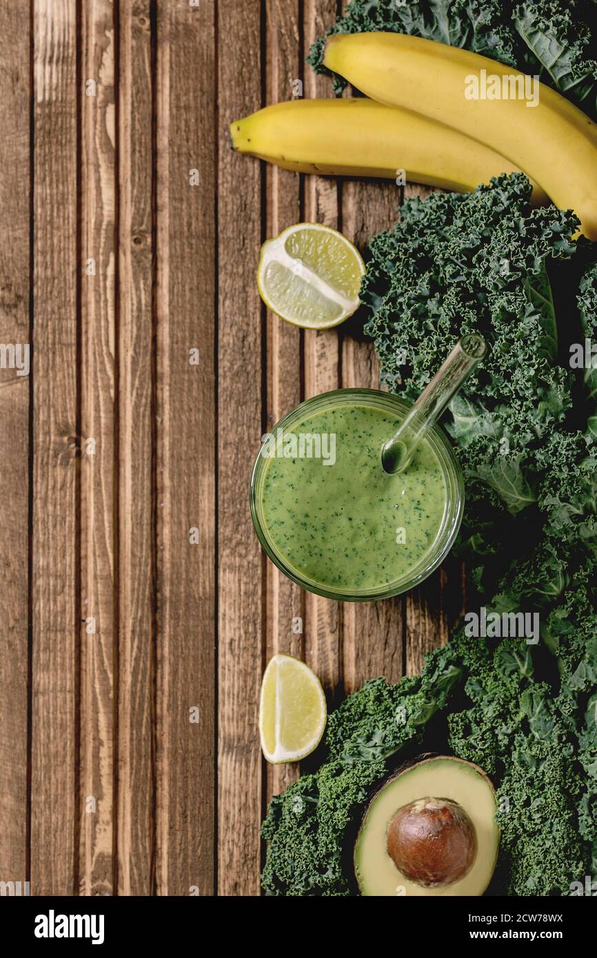 Verre de smoothie végétalienne saine et verte, paille de verre. Ingrédients ci-dessus. Kale, bananes, avocat, chaux, sur fond de planche en bois. Plat, espace Banque D'Images