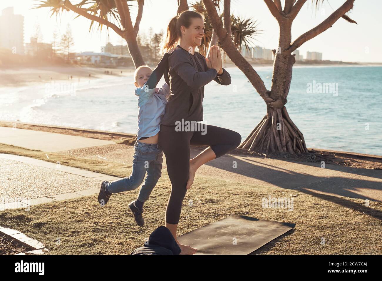 Jeune mère et fils faisant de la gymnastique et s'étirant dans le parc de la ville sur le soleil avec vue sur l'océan. Femme en yoga asana et garçon sur ses épaules Banque D'Images