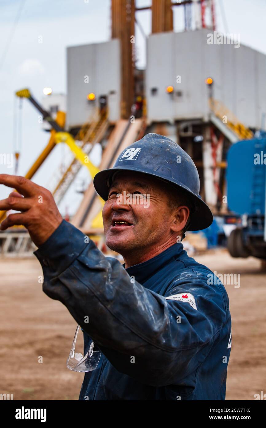 Ouvrier du pétrole asiatique adulte en vêtements de travail bleus et casque de sécurité Avec logo BJ (Baker Hughes) Poser et sourire sur l'engin de forage de pétrole bckgd.Zhaik-Munai gisement d'huile Banque D'Images