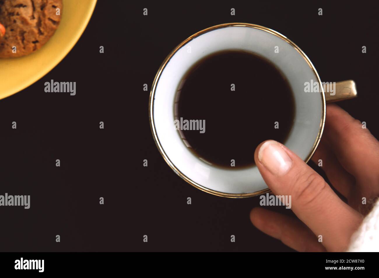 La main de la femme tient une tasse de café en porcelaine dorée sur fond noir. Assiette jaune avec biscuits aromatisés et gouttes de couleur Banque D'Images