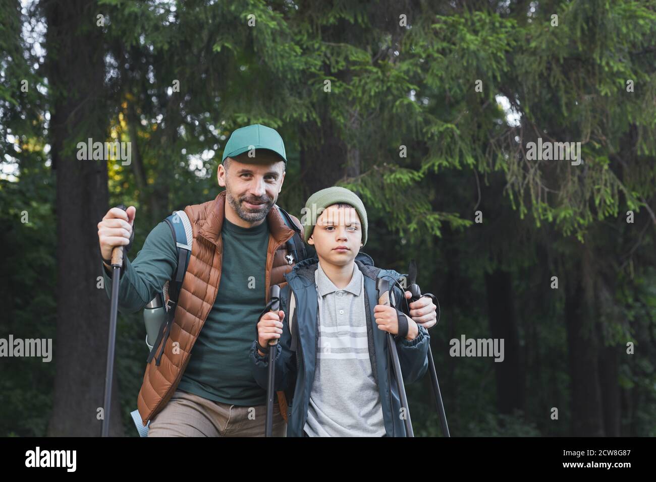 Portrait à la taille de père heureux et fils randonnée ensemble et regardant l'appareil photo tout en marchant dans la forêt avec des bâtons de randonnée, espace de copie Banque D'Images