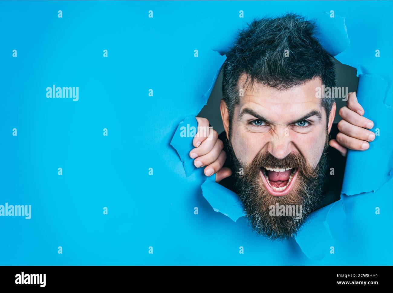 Homme barbu qui fait trou dans le papier. Vue de la face mâle en colère à travers le trou du papier. Homme en colère regardant à travers le trou. Homme barbu en papier. Émotions. Banque D'Images