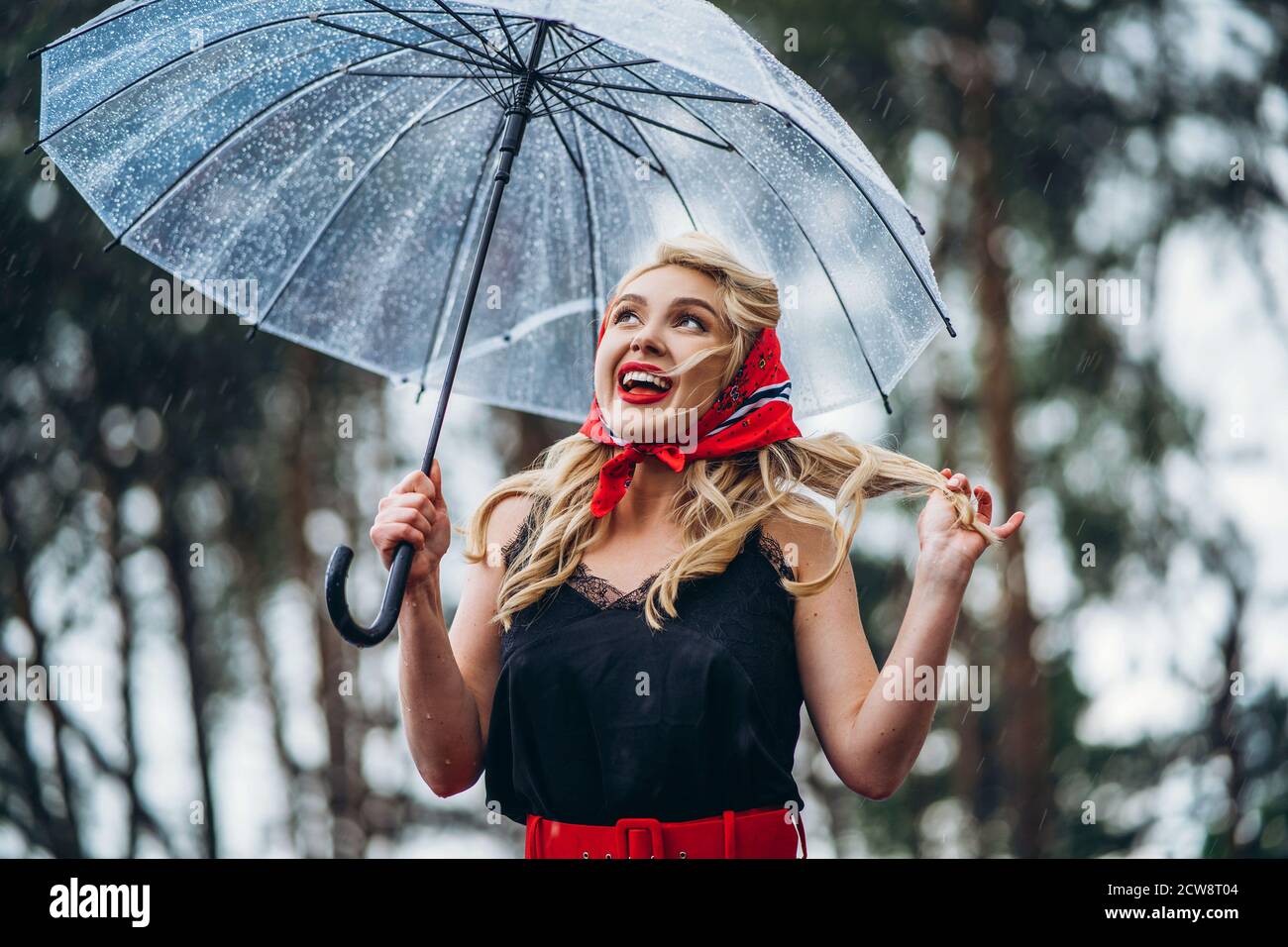 Joyeux blond stylé avec des lèvres rouges qui marchent jour de pluie à l'extérieur avec parapluie transparent Banque D'Images
