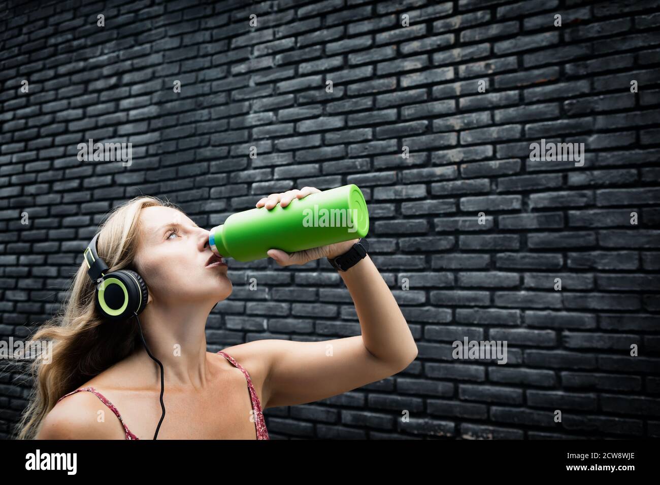 Jeune femme blonde sur fond noir buvez de l'eau de source fraîche provenant d'un flacon vert réutilisable. Un style de vie sain en ville d'été. Banque D'Images