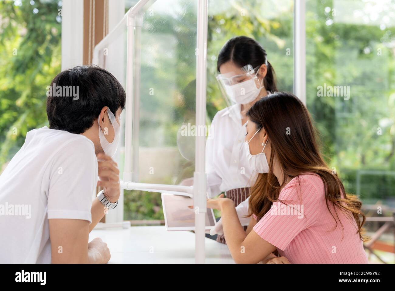 La serveuse asiatique porte un masque facial et un masque facial en utilisant une tablette pour montrer le menu électronique du restaurant et recommande aux clients de couple. Nouvelle technologie normale Banque D'Images