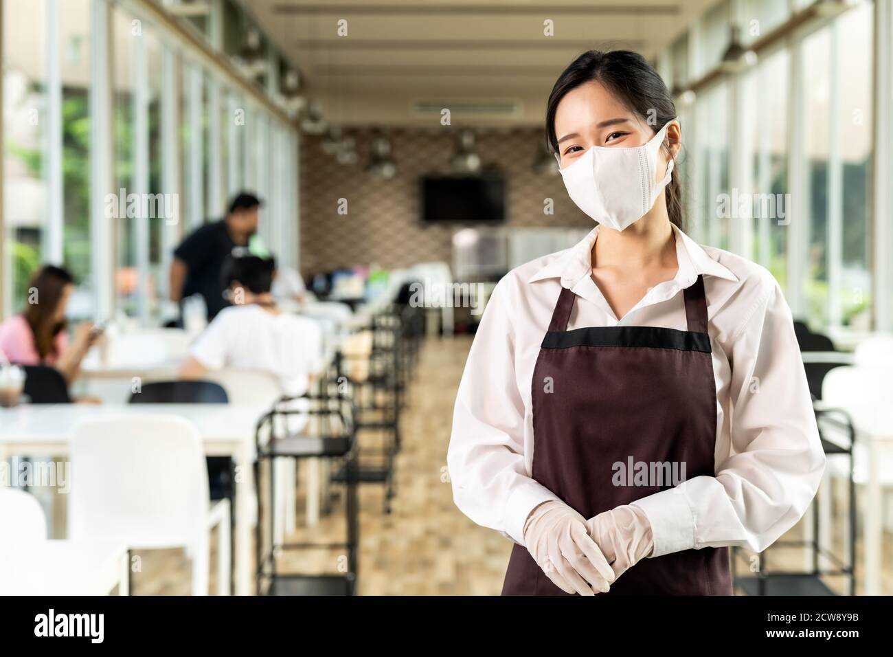 Portrait élégant asiatique porte-serveuse visage avec le fond des clients assis dans la table de social distance dans le restaurant à l'intérieur. Nouveau restaurant normal lif Banque D'Images