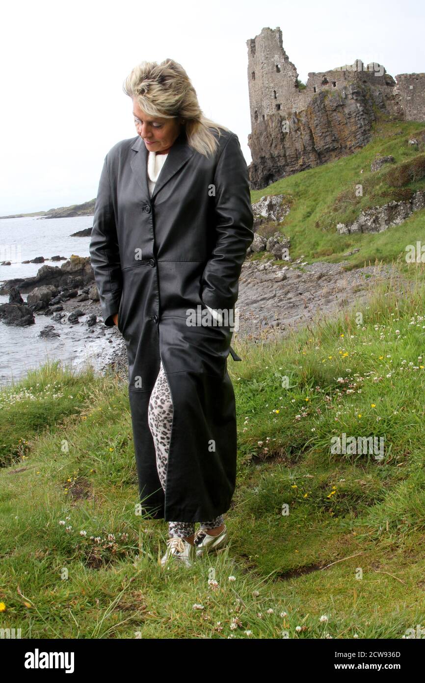 Femme de race blanche mature avec une coupe blonde de mulet en long manteau  de cuir et voir à travers unbrella regardant autour des ruines du château  et vieux village de pêcheurs