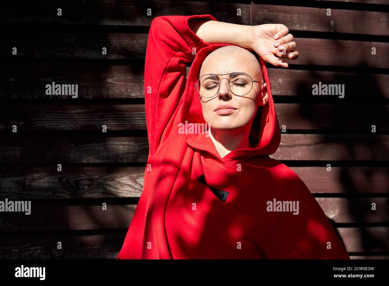 Femme chauve fraîche portant un sweat à capuche rouge et des lunettes debout sur fond de bois. Banque D'Images