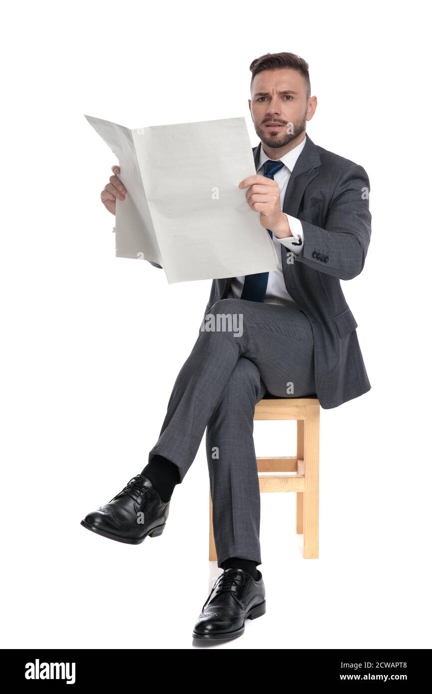 un jeune homme d'affaires confus lisant des journaux, assis isolé sur fond blanc, plein corps Banque D'Images