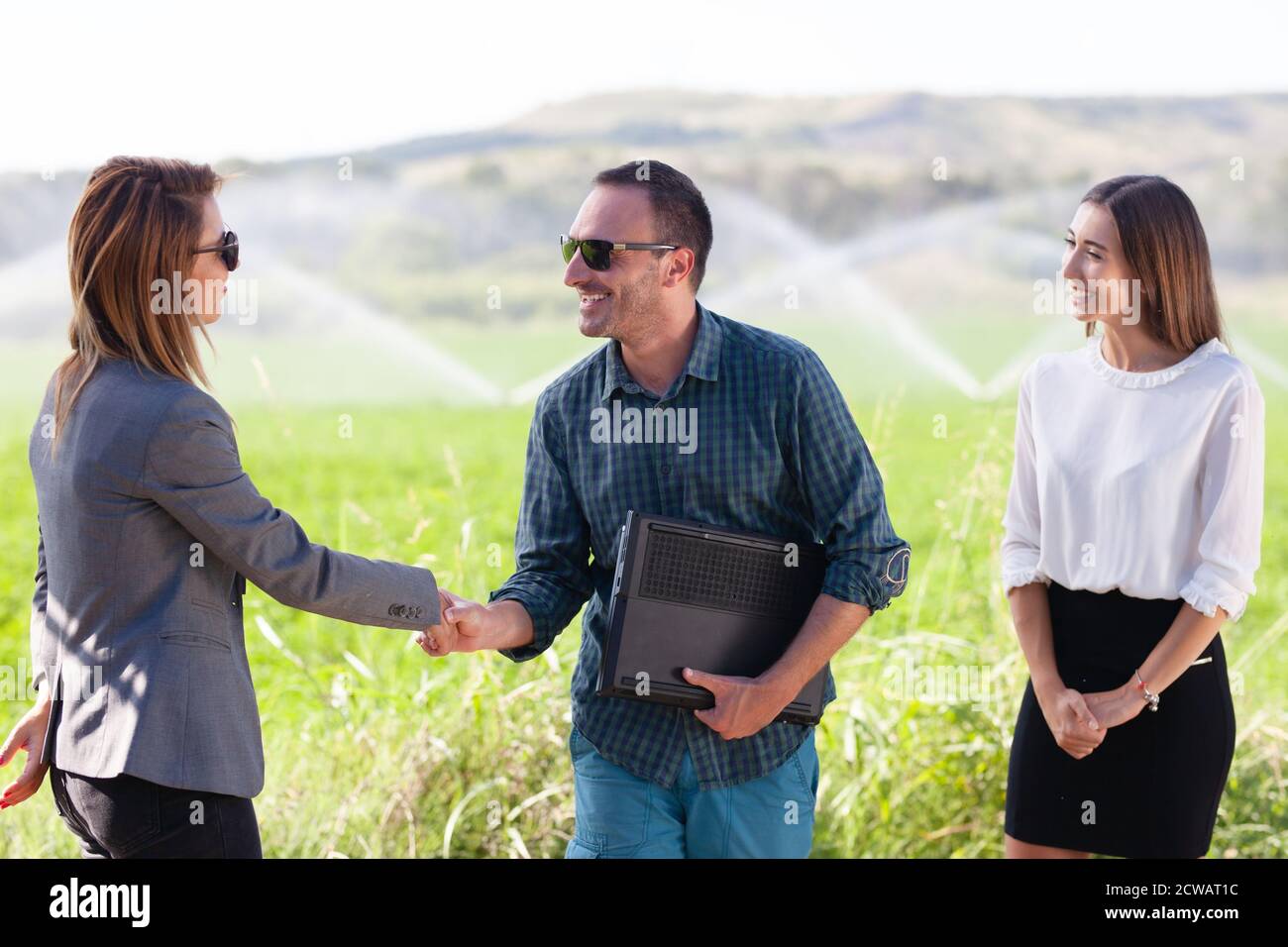 Les hommes d'affaires se secouent la main dans un champ vert avec un système d'irrigation agricole. Arroseurs d'eau en arrière-plan. Banque D'Images
