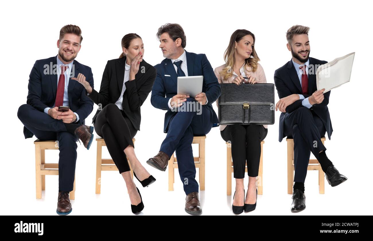 Équipe de 5 hommes d'affaires tenant, téléphone, tablette, journal et porte-documents, discutant les uns avec les autres en attendant un entretien d'emploi assis sur des chaises sur W Banque D'Images