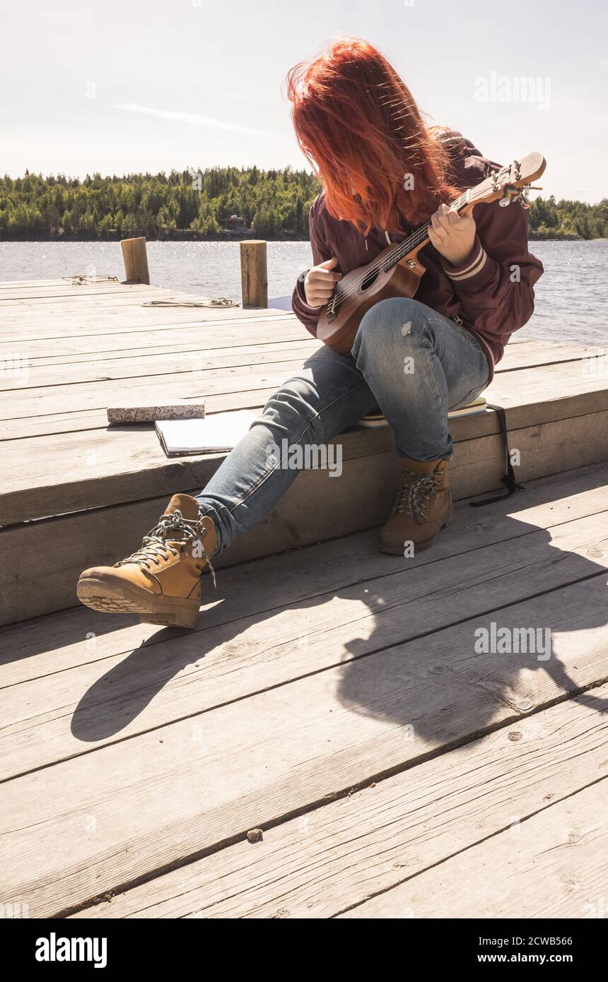 Une jeune fille aux cheveux rouges chante une chanson avec ukulele assis sur une jetée en bois par beau temps Banque D'Images