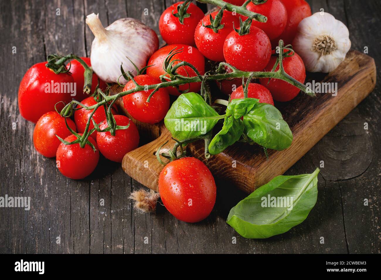 Assortiment de tomates cerises colorées avec ail et basi sur petite planche à découper sur une ancienne table en bois. Banque D'Images