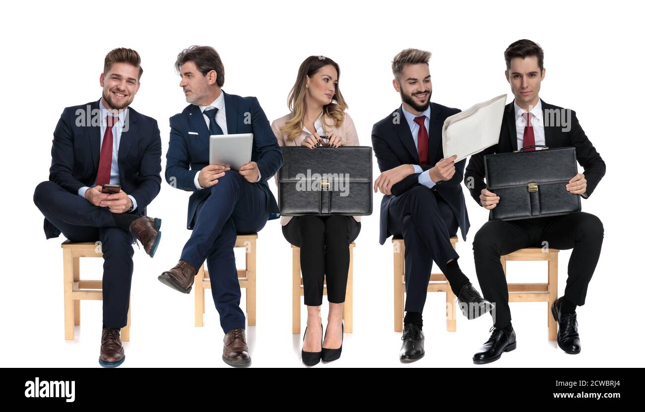 Équipe de 5 hommes d'affaires tenant, téléphone, tablette, journal et porte-documents tout en attendant un entretien d'emploi assis sur des chaises sur fond blanc de studio Banque D'Images