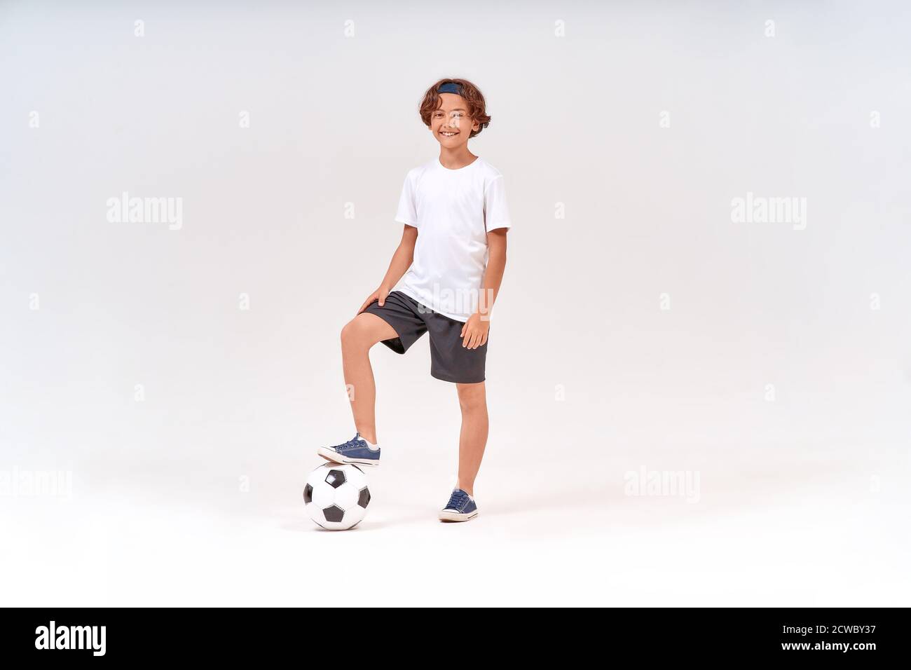 Petit joueur. Prise de vue en longueur d'un adolescent heureux avec ballon de football regardant l'appareil photo et souriant tout en se tenant isolé sur fond gris Banque D'Images