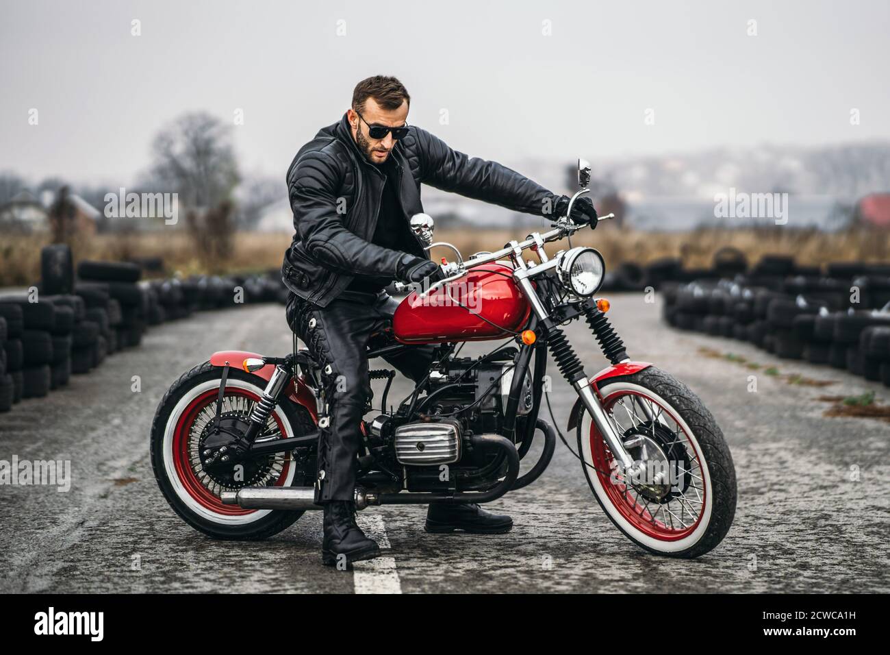 Moto rouge avec pilote. Un homme dans une veste et un pantalon en cuir noir  se tient latéralement au milieu de la route. Les pneus sont posés sur  l'arrière-plan Photo Stock -