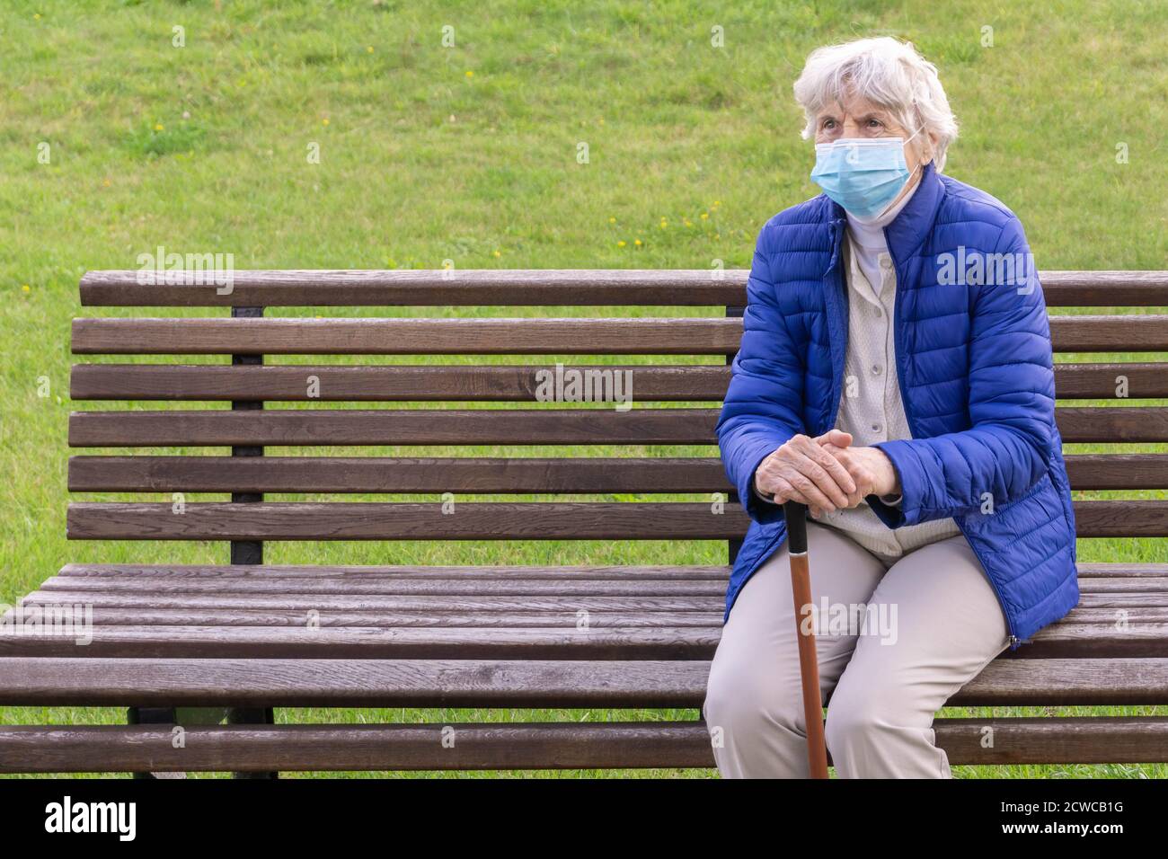 Femme âgée portant un masque médical assise sur un banc à l'extérieur et tenant un bâton de marche. Femme aux cheveux gris avec masque de protection sur la canne sur le banc Banque D'Images