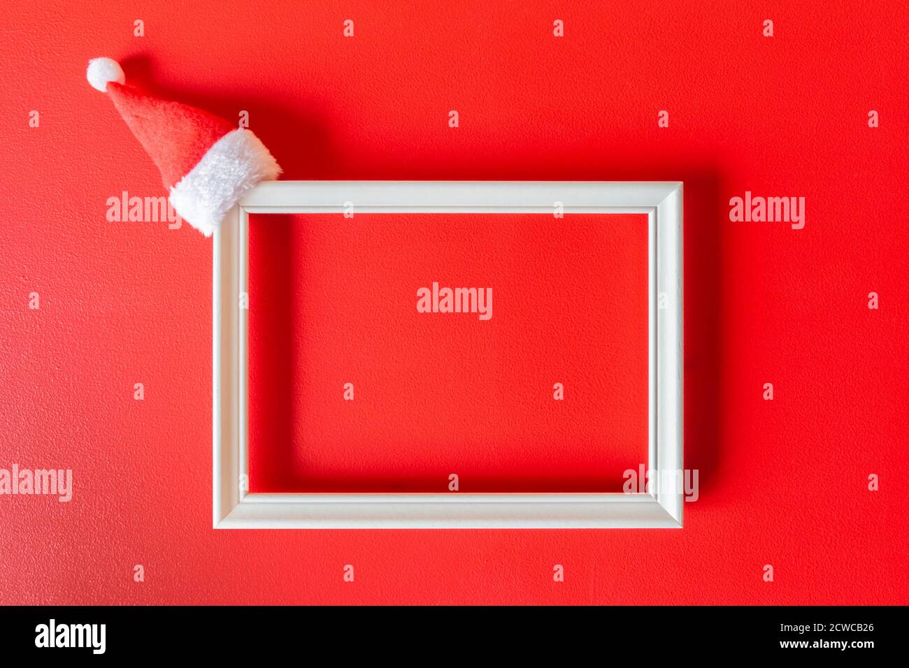 Cadre blanc avec petit chapeau de Père Noël sur fond peint en rouge. Décoration de fête minimaliste avec espace pour les copies. Maquette pour Noël. Noël Banque D'Images