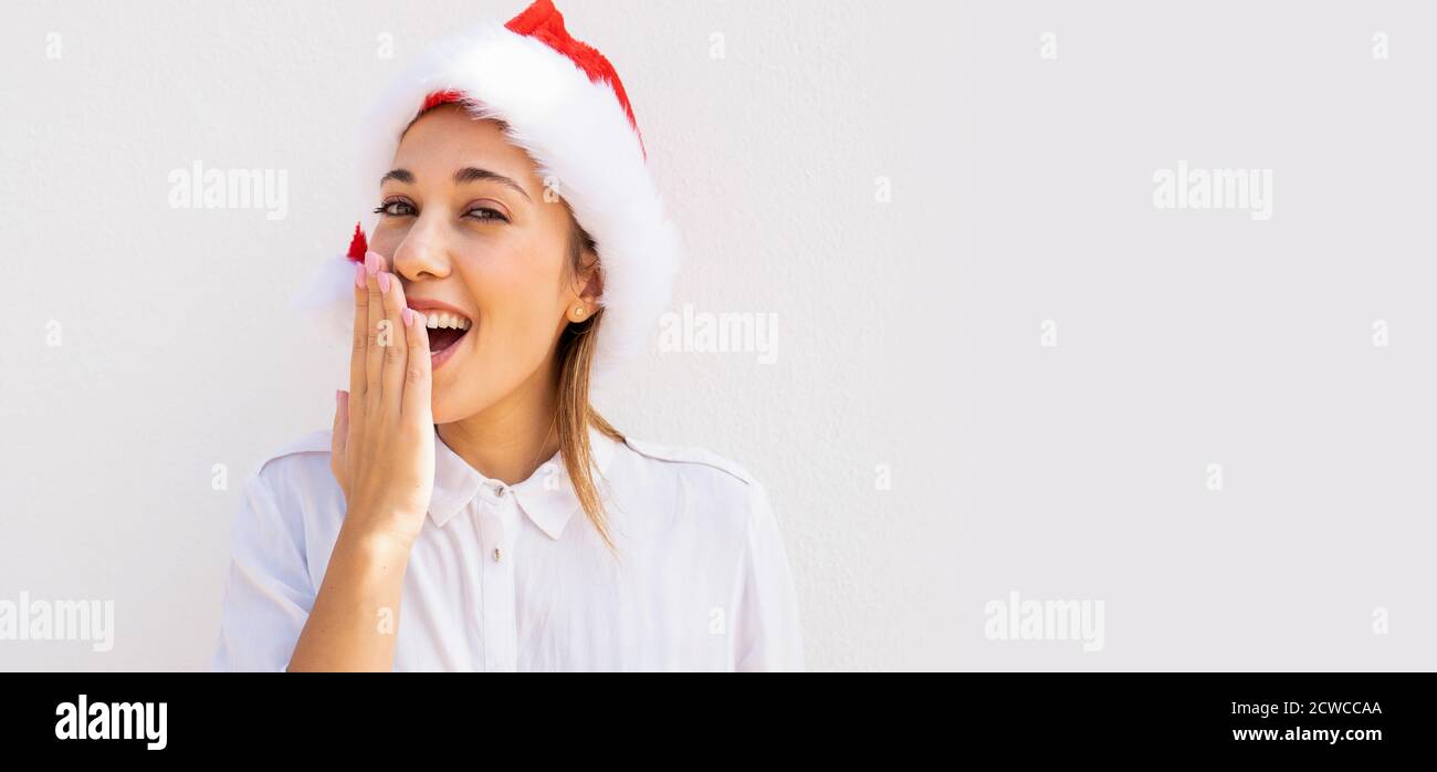 Jeune belle femme blonde caucasienne avec un chapeau de Père Noël surprenant avec sa main devant sa bouche avec grand arrière-plan d'espace de copie blanc - Banque D'Images