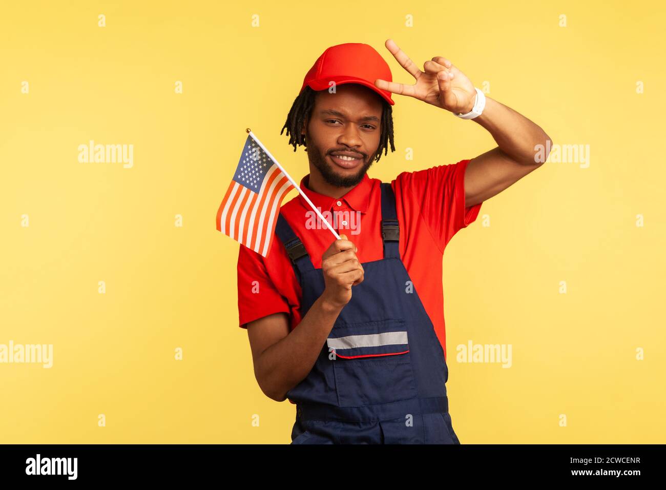 Travailleur afro-américain en uniforme avec barbe et dreadlocks tenant le drapeau américain et montrant le geste de paix avec les doigts regardant la caméra. Intérieur s Banque D'Images