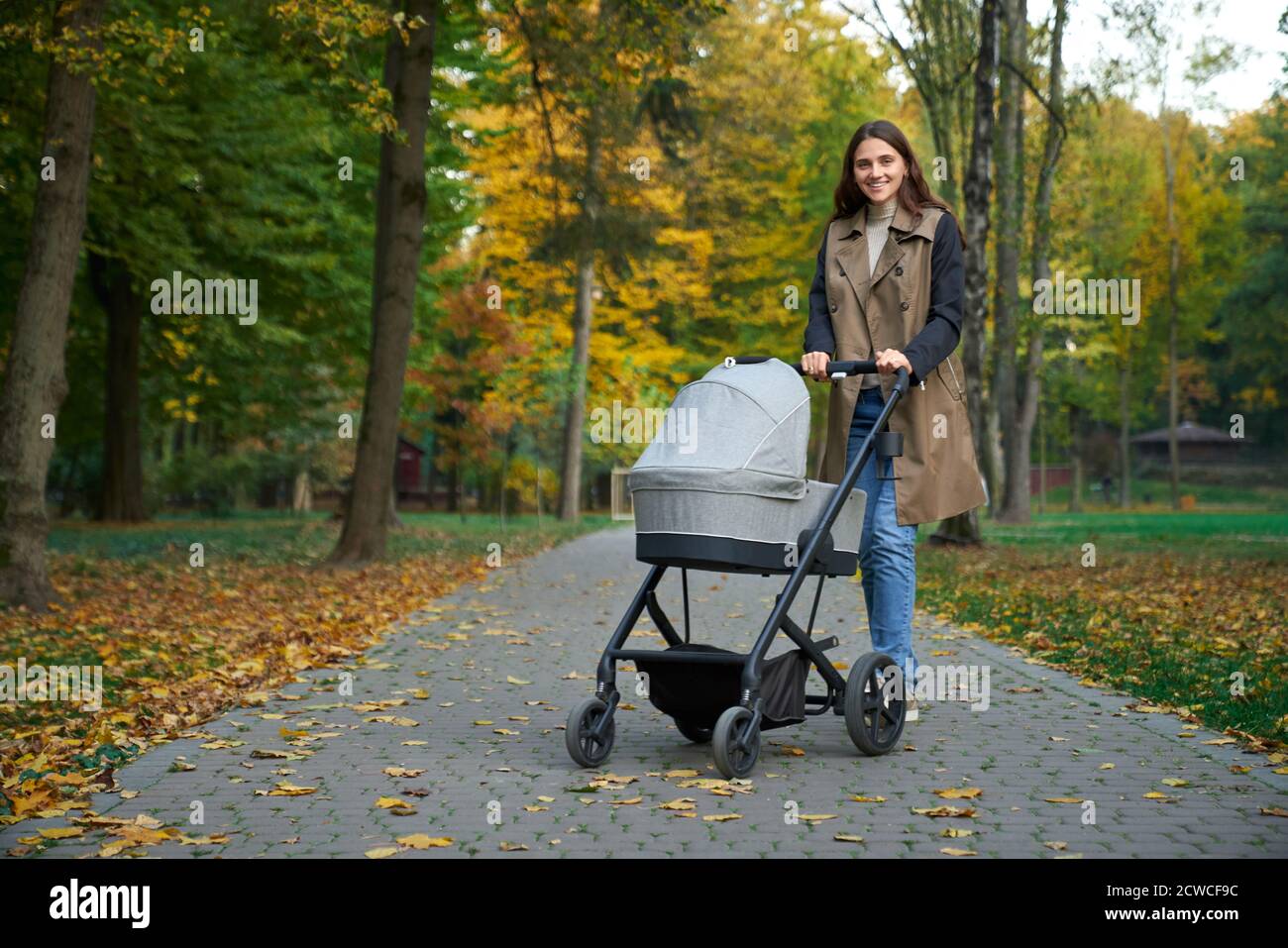 Vue latérale d'une femme dans un long manteau debout avec une poussette dans le parc d'automne. Mère avec bébé souriant à l'appareil photo. Concept de poussette pour bébé. Banque D'Images