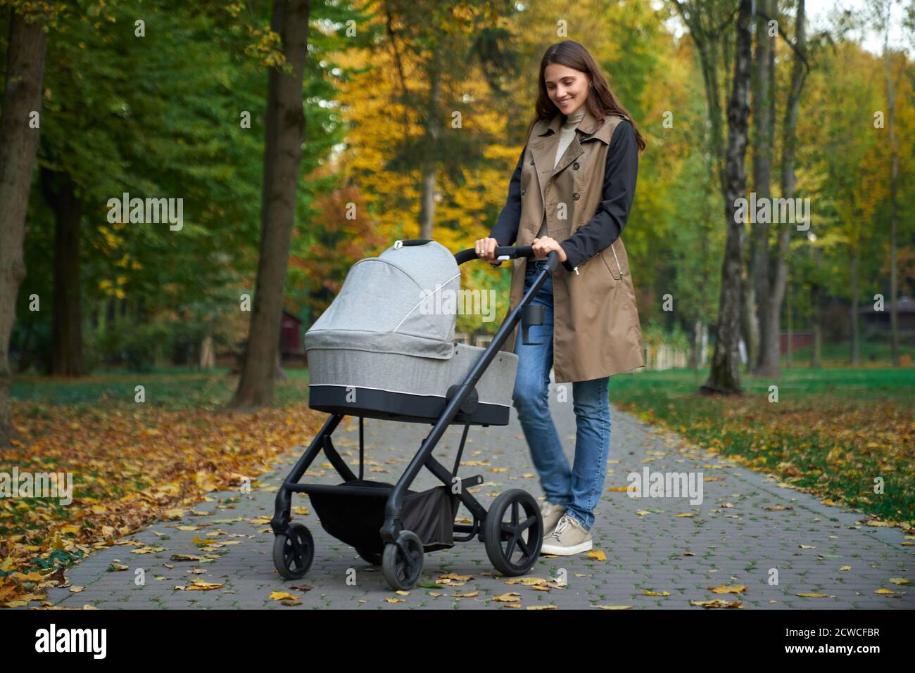 Vue latérale d'une femme mince pleine hauteur en manteau avec poussette grise. Une fille se tient dans le parc et regarde le bébé dans un chariot gris. Concept de pose. Banque D'Images
