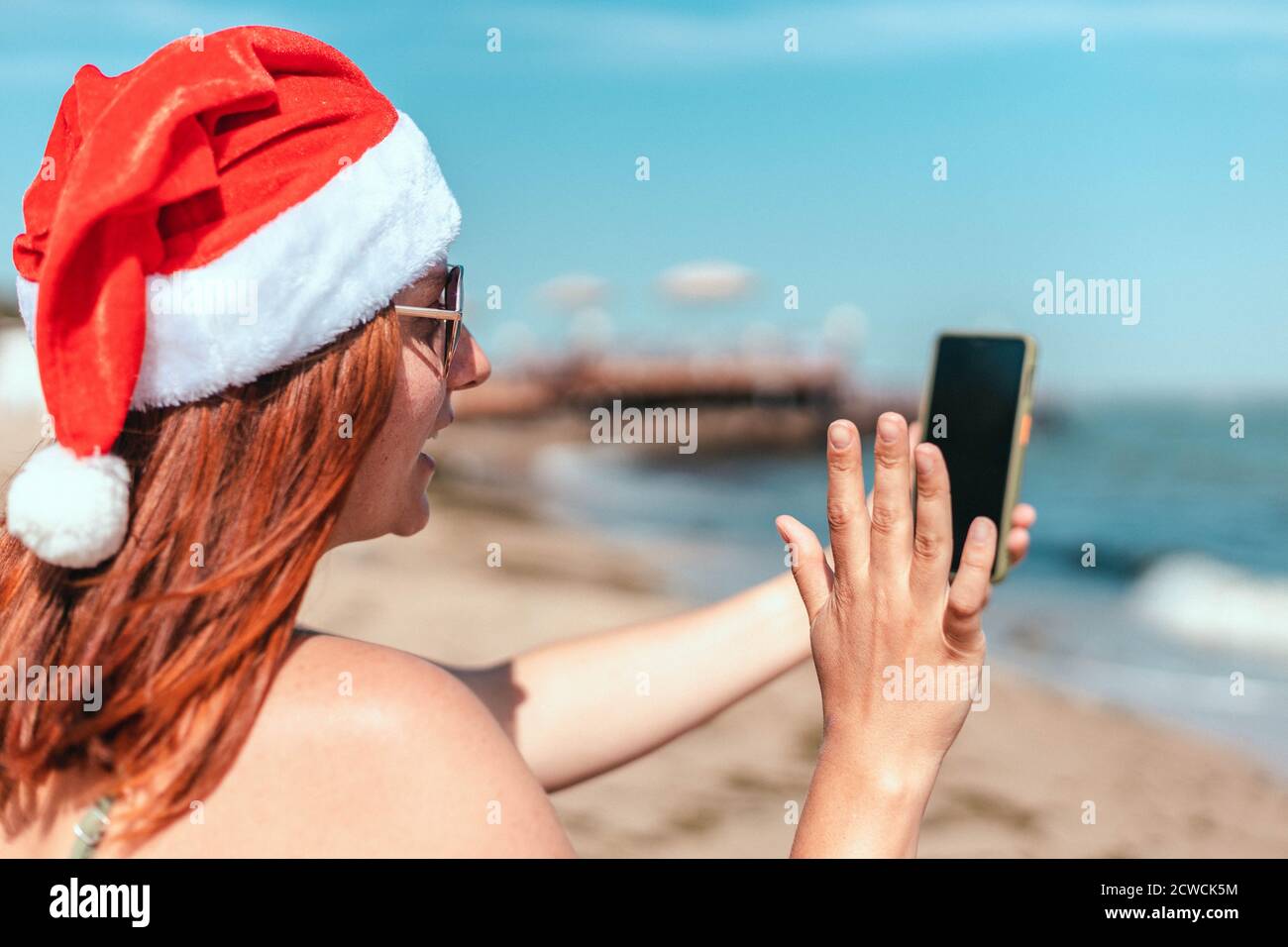 Jeune belle fille en chapeau et maillot de bain du père noël rouge fait un selfie sur le bord de mer Banque D'Images