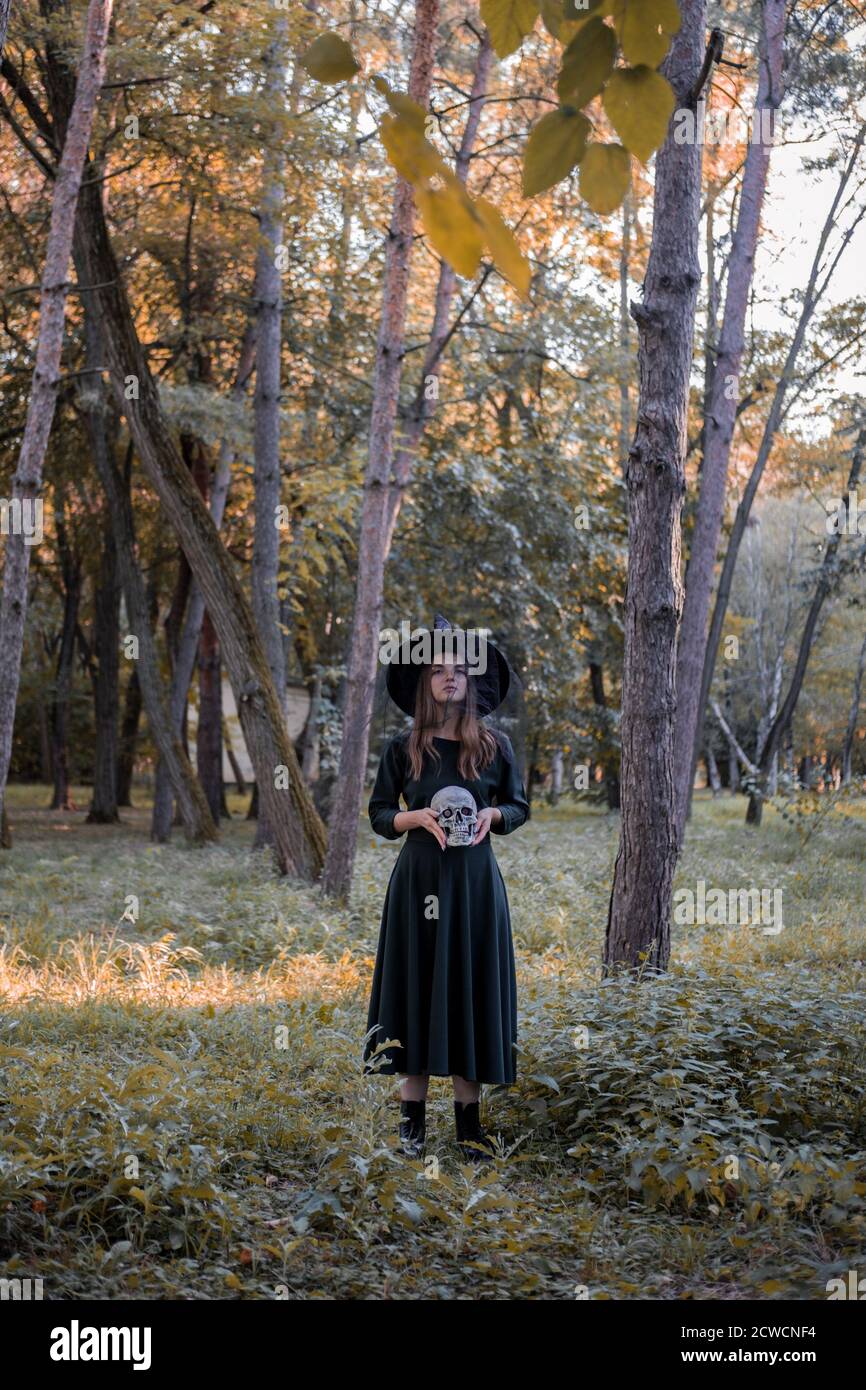 Jeune belle femme mignonne en robe sombre et chapeau de sorcière tient un crâne dans ses mains. Costume de fête d'Halloween. Forêt, parc avec arbres d'automne Banque D'Images