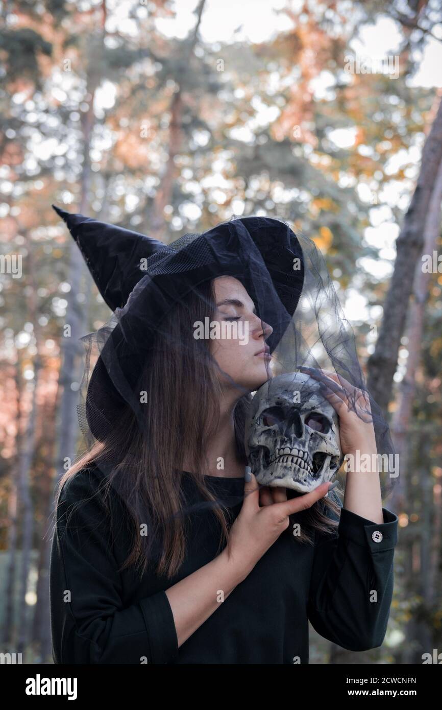 Jeune belle femme mignonne en robe sombre et chapeau de sorcière tient un crâne dans ses mains. Costume de fête d'Halloween. Forêt, parc avec arbres d'automne Banque D'Images