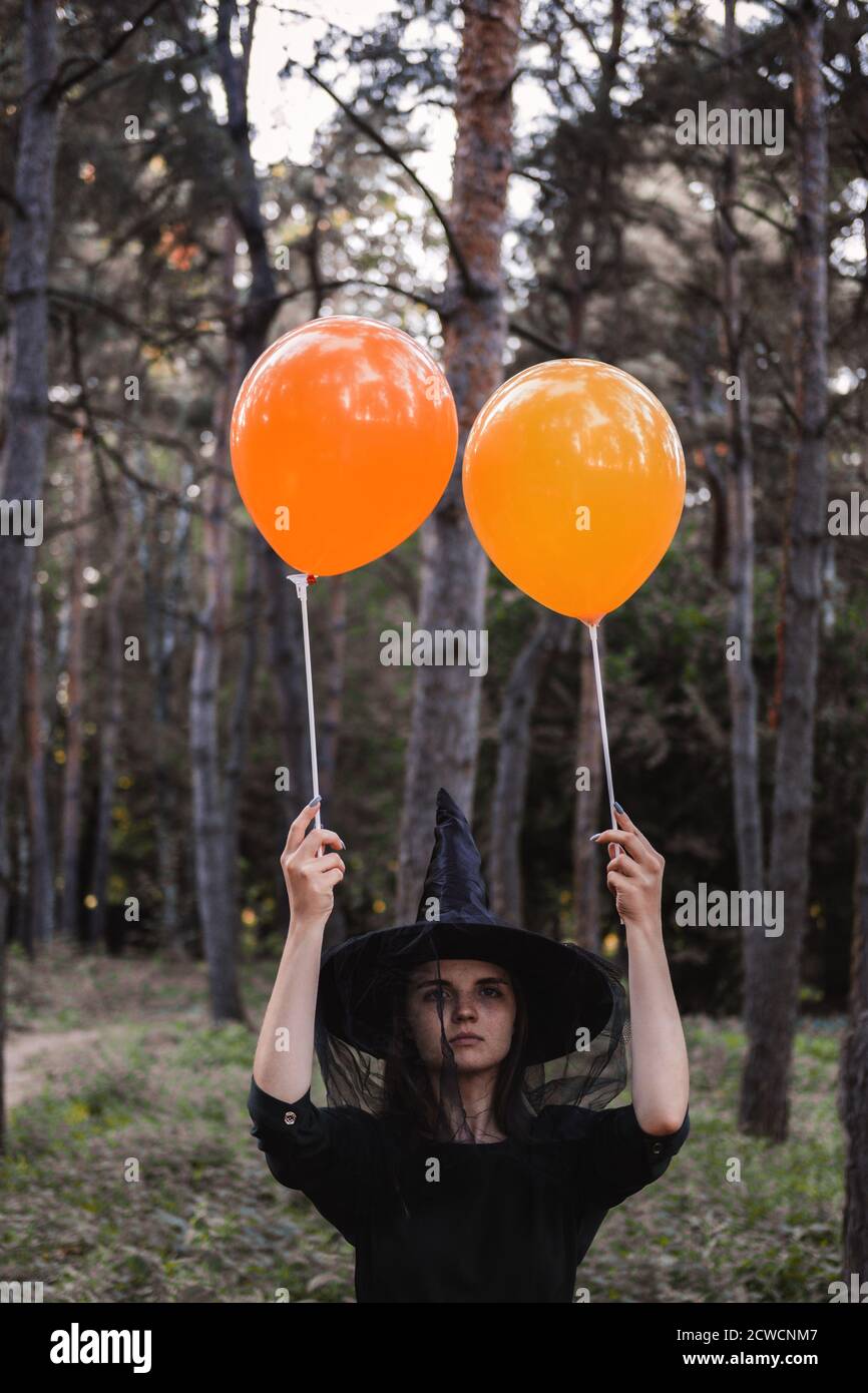 Jeune mignonne belle femme en robe sombre et chapeau de sorcière tient des ballons d'orange dans ses mains. Costume de fête d'Halloween. Forêt, parc avec arbres d'automne Banque D'Images