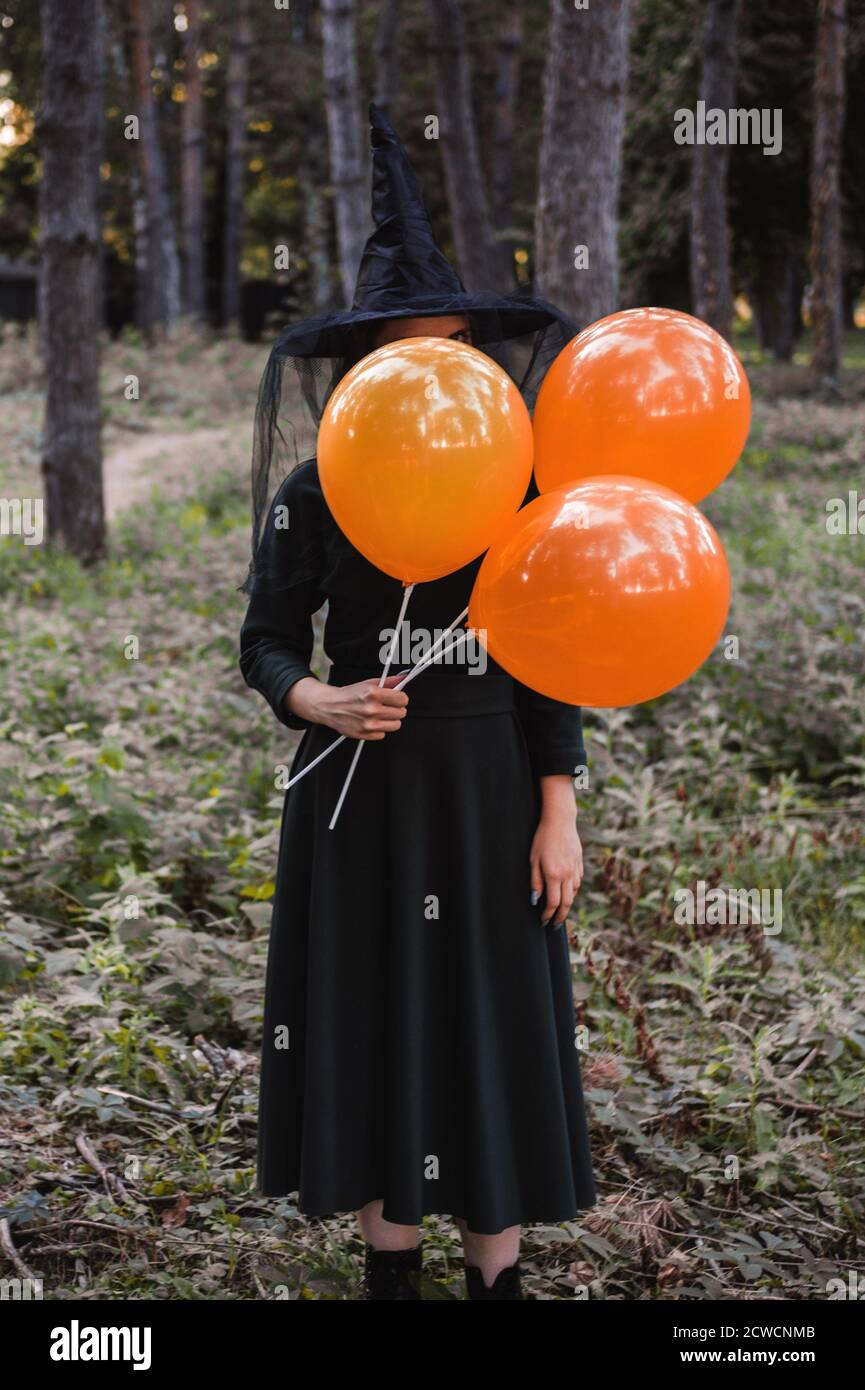 Jeune mignonne belle femme en robe sombre et chapeau de sorcière tient des ballons d'orange dans ses mains. Costume de fête d'Halloween. Forêt, parc avec arbres d'automne Banque D'Images