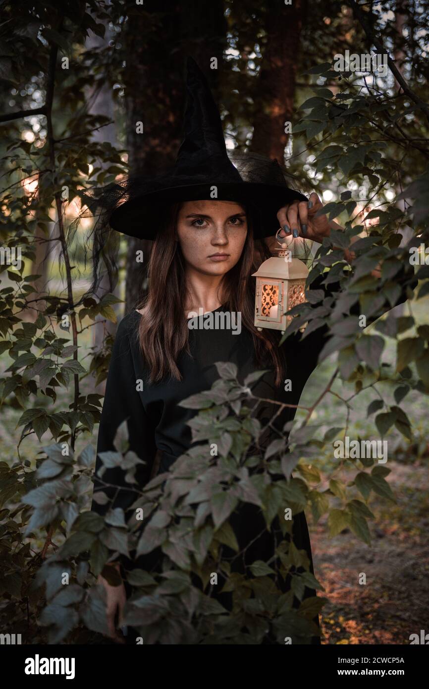 Une jeune femme en robe sombre et chapeau de sorcière tient une lanterne avec des bougies dans ses mains et illumine la forêt. Costume de fête d'Halloween. Garez-vous avec l'autom Banque D'Images