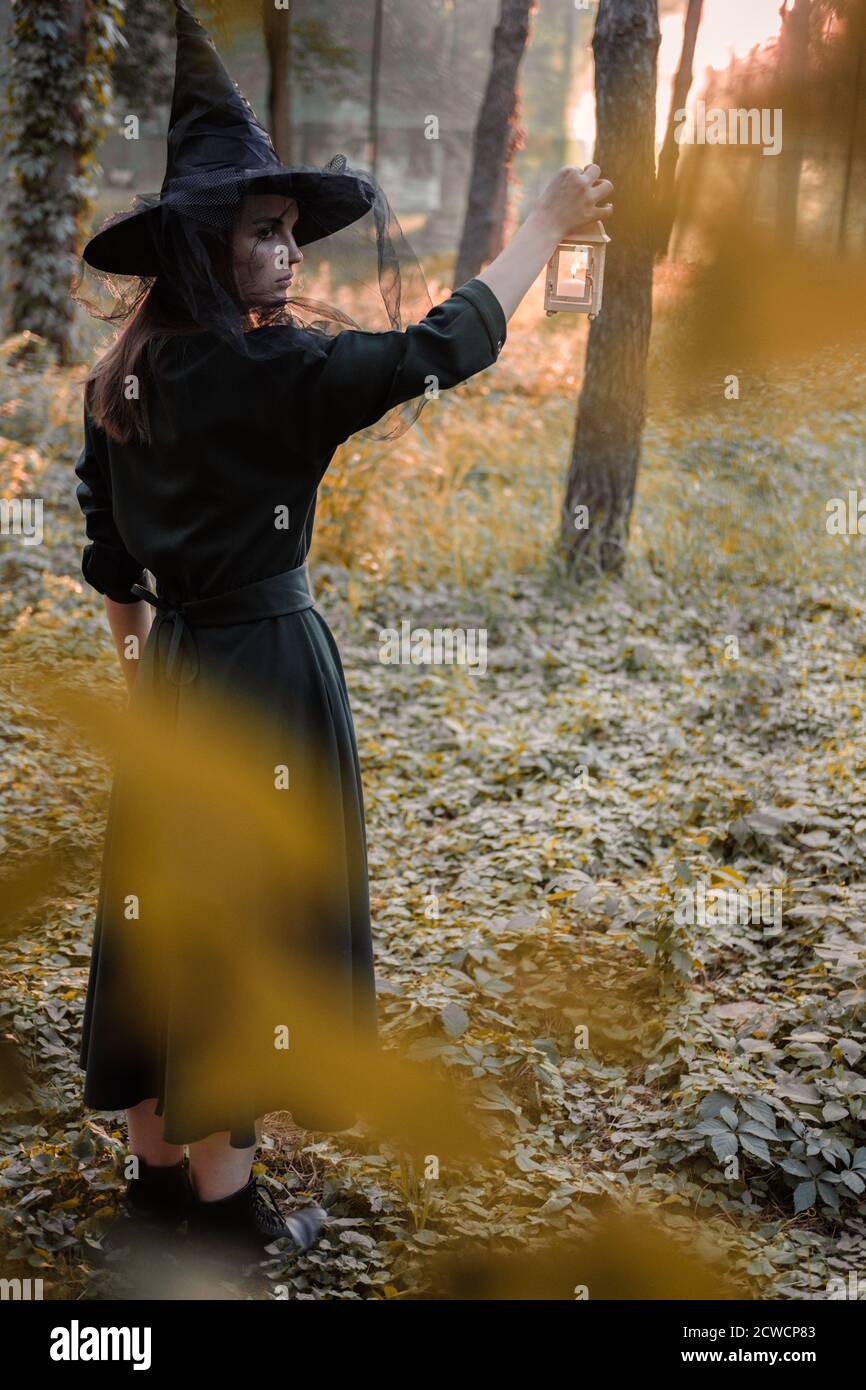 Une jeune femme en robe sombre et chapeau de sorcière tient une lanterne avec des bougies dans ses mains et illumine la forêt. Costume de fête d'Halloween. Garez-vous avec l'autom Banque D'Images