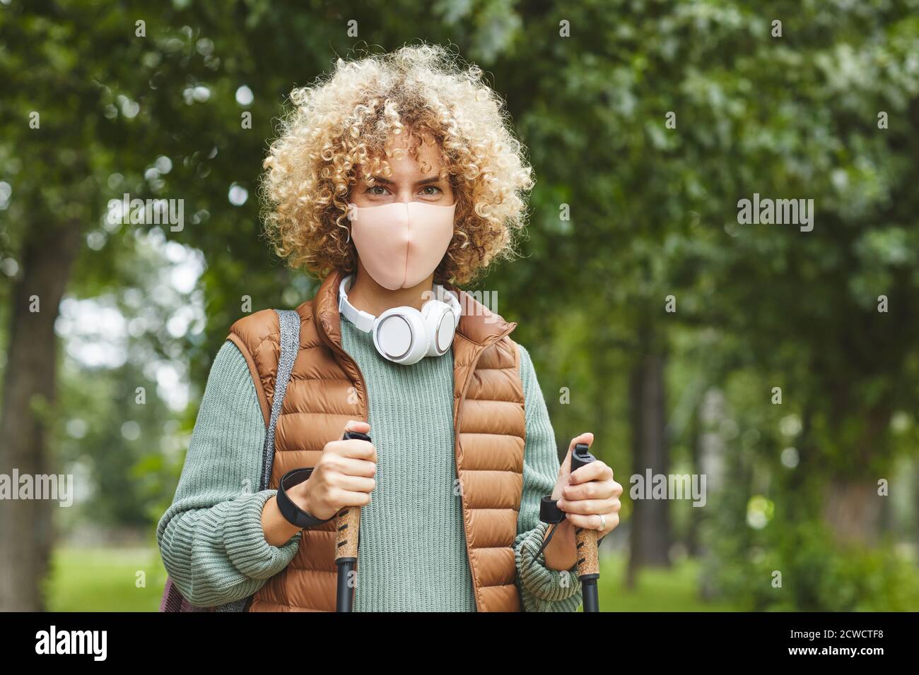Portrait d'une jeune femme avec des cheveux bouclés portant un masque de protection regarder la caméra pendant la marche sportive dans le parc Banque D'Images