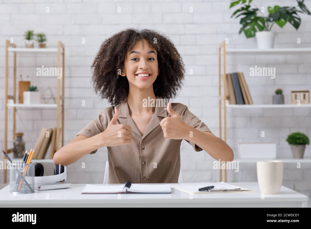 Moderne maison blogger, grand examen et passe-temps. Femme afro-américaine souriante montrant les pouces et regardant l'appareil photo, assis à une table avec un ordinateur portable Banque D'Images