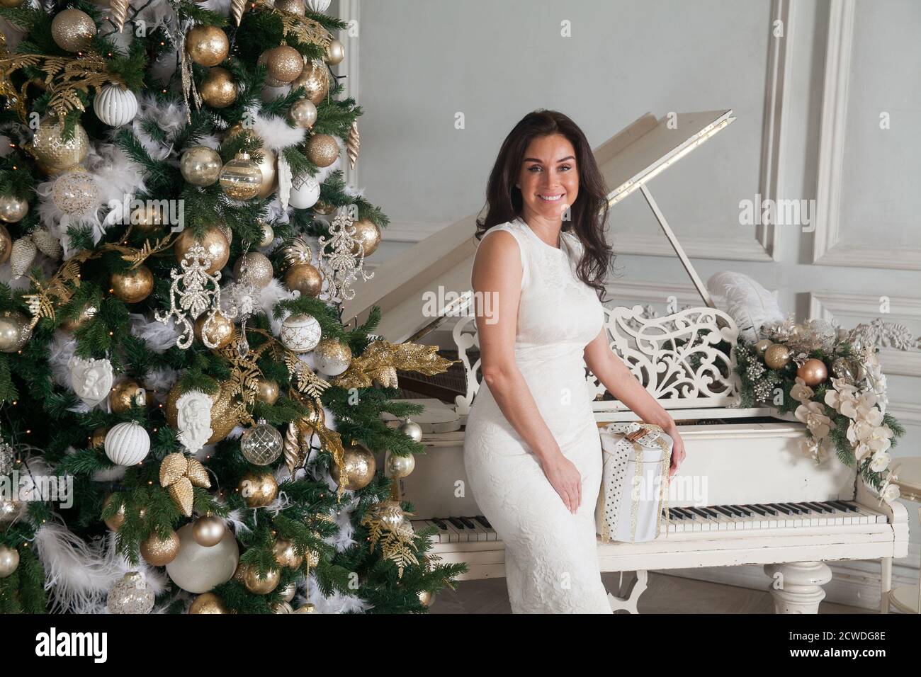 Joyeux Noël et joyeuses fêtes. Belle jeune femme brune en robe blanche de  soirée à cocktail posant avec un piano blanc et décorée Noël Photo Stock -  Alamy