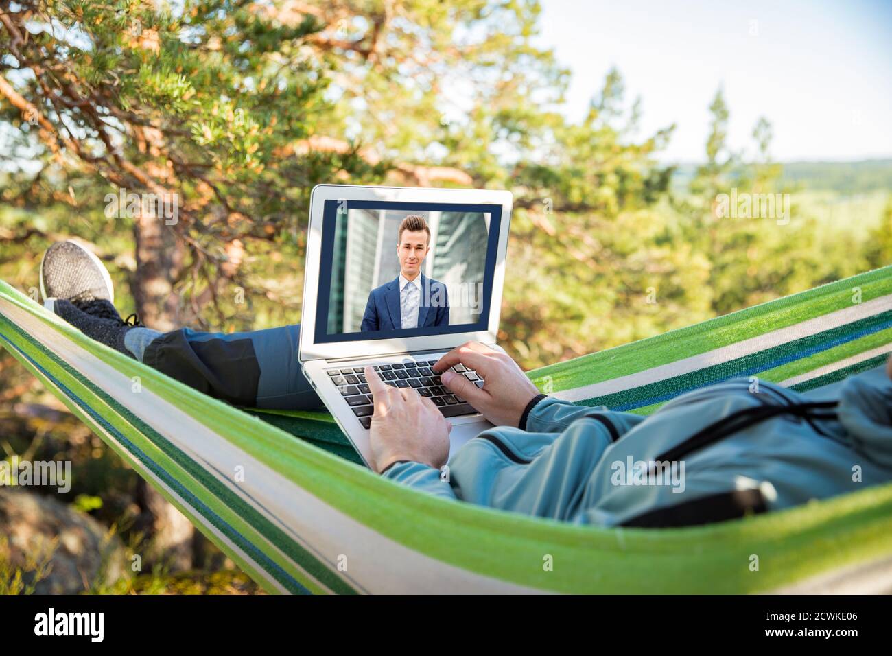 Un homme qui travaille sur un ordinateur portable tout en étant allongé dans un hamac dans les bois. Auto-isolation, freelance, travail à distance et distanciation. Appel vidéo avec des collègues. Banque D'Images