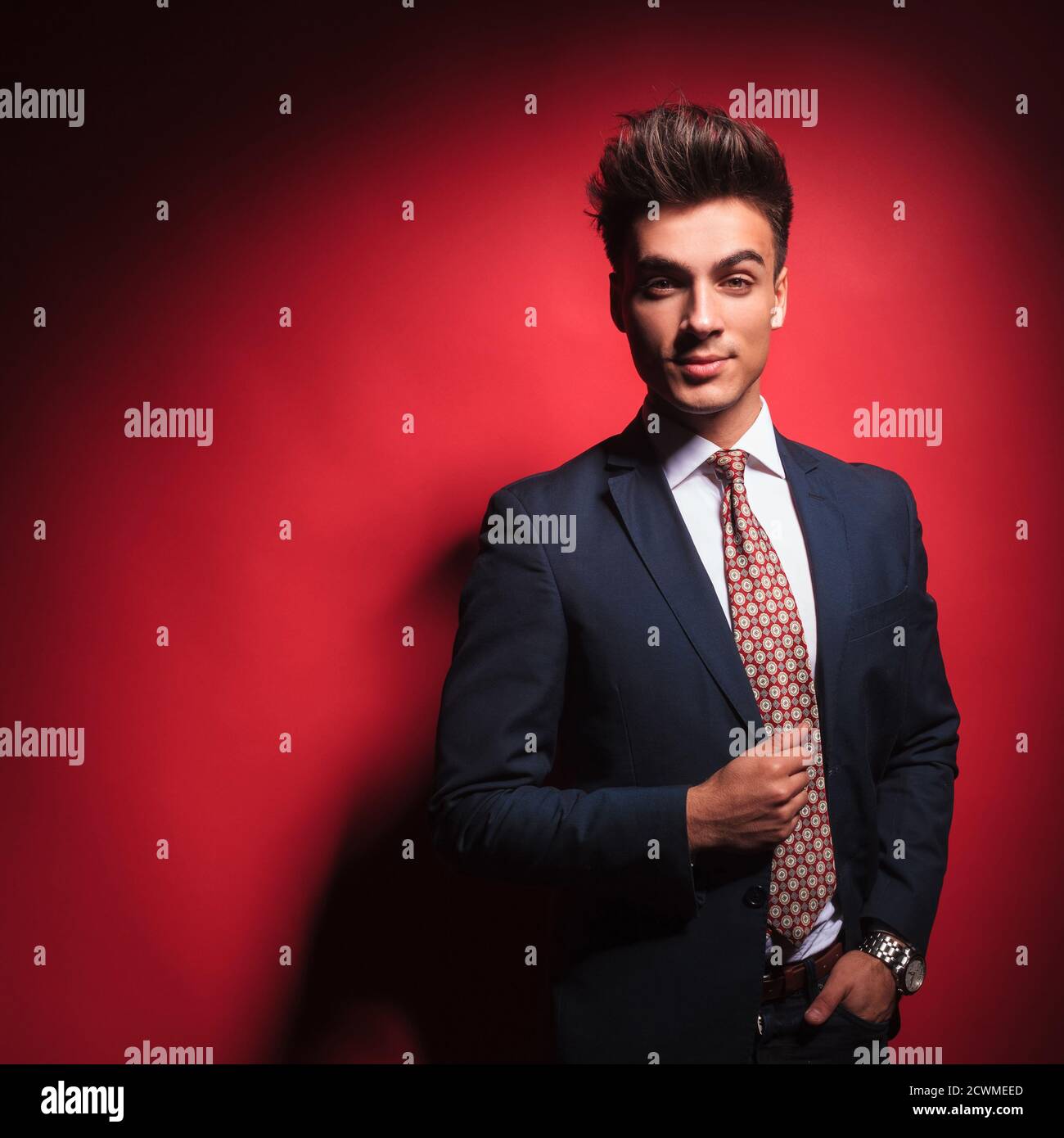 portrait de jeune homme d'affaires attrayant en costume noir avec cravate  rouge, posant avec la main dans la poche, l'organisation de sa veste, dans  le fond rouge studio pendant Photo Stock -