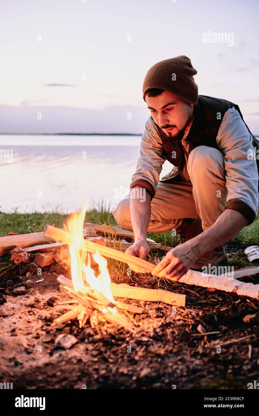 Jeune randonneur barbu sérieux dans le chapeau mettant du bois pendant la mise en place feu près de la côte au coucher du soleil Banque D'Images