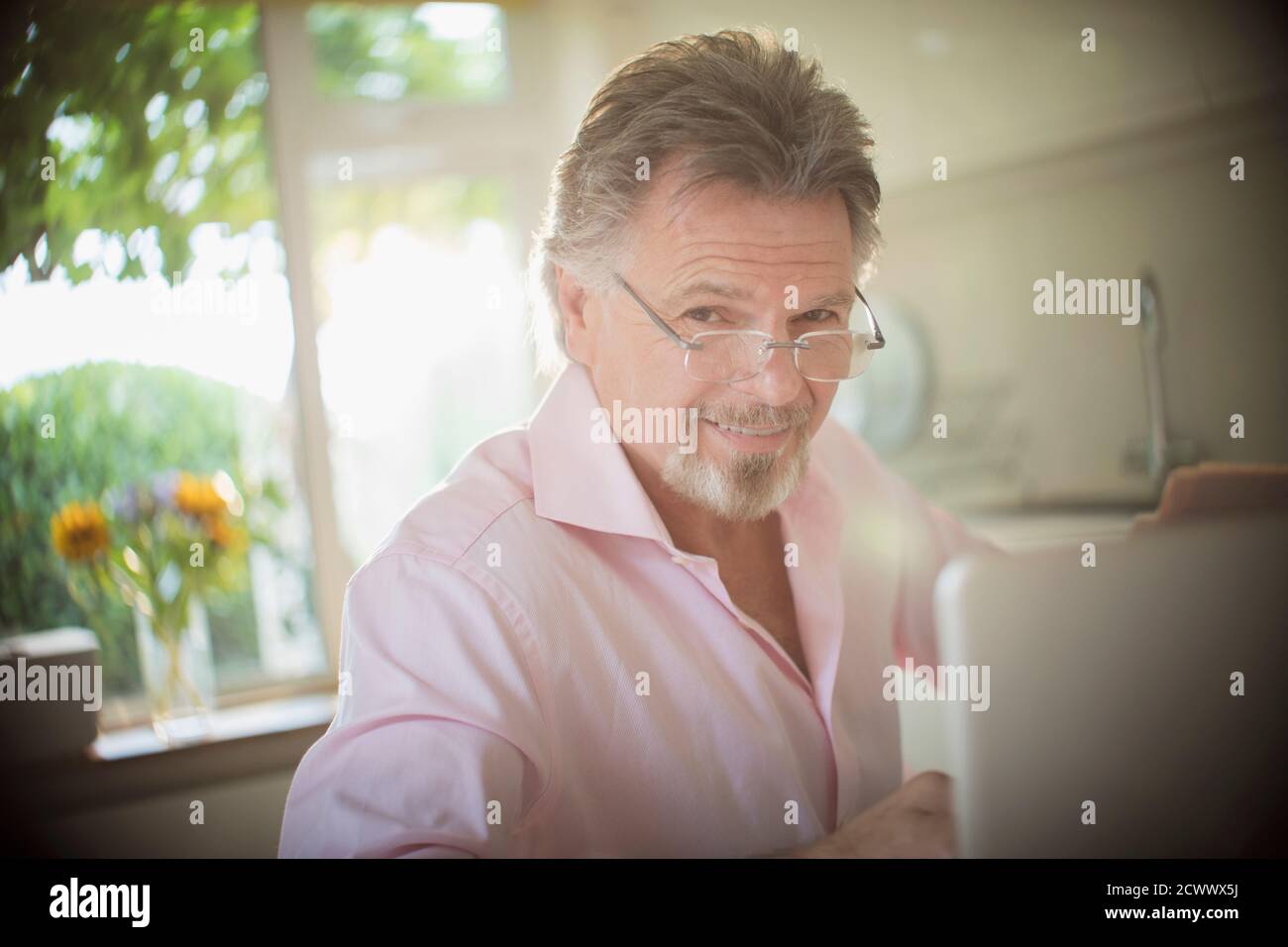 Portrait souriant homme senior travaillant à l'ordinateur portable dans la cuisine ensoleillée Banque D'Images